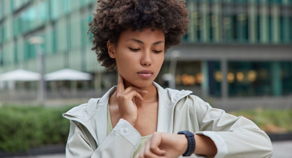 Fitness lady counts hearbeat checks pulse on neck concentrated at smartwatch monitors her health after workout dressed in sportswear poses outside against modern city building. Sport and health