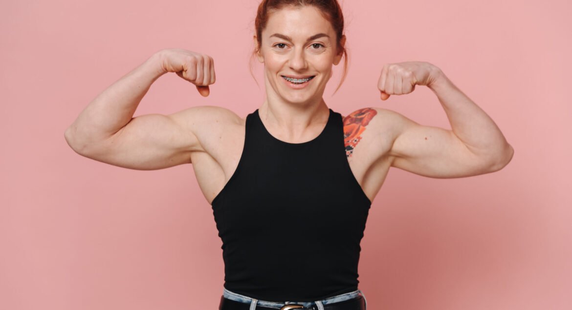 Happy sporty muscular woman with red hair in t-shirt and jeans smiling with braces and showing biceps
