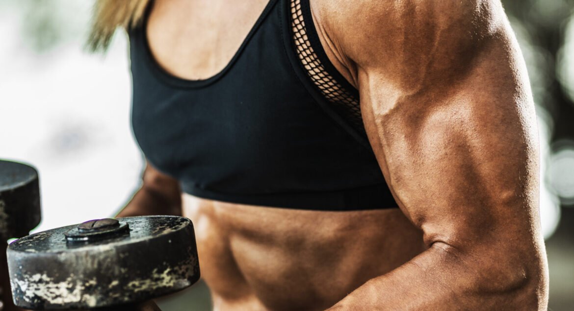Close-up of a muscular woman bodybuilder doing biceps exercises with dumbbells in the nature.