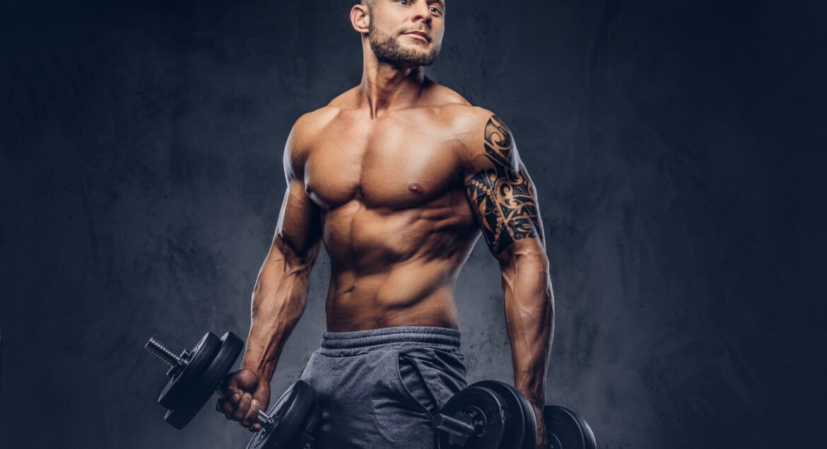 Powerful stylish bodybuilder with tattoo on his arm, doing the exercises with dumbbells. Isolated on a dark background.